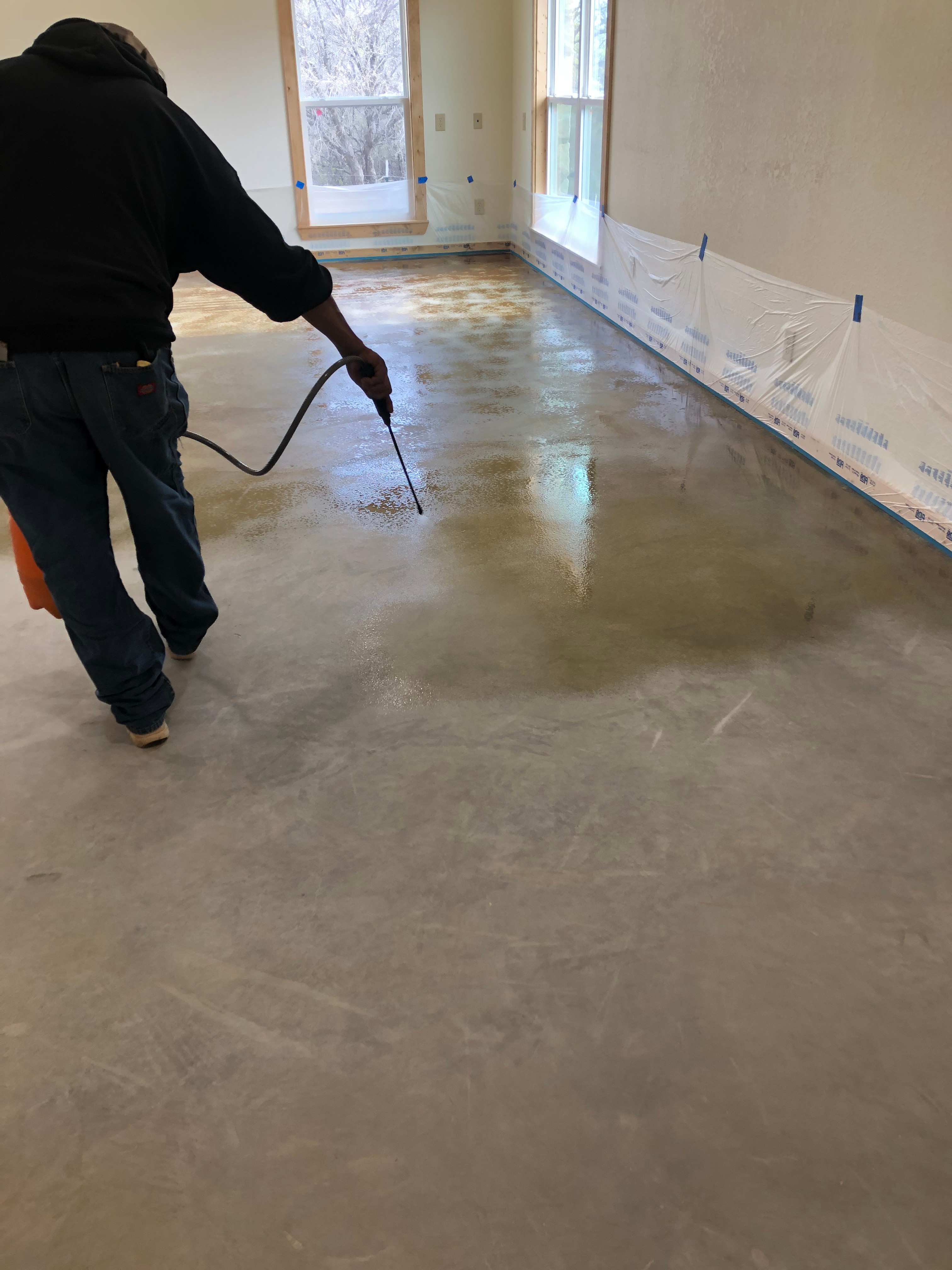 Before: R.G. Pro Floors technician applying acid stain to bare concrete floor with spray applicator, walls protected with plastic sheeting in Brownwood TX